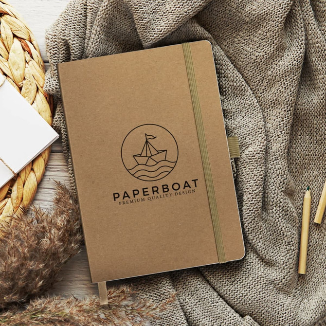 A brown notebook with a logo, lying on a textured fabric beside pens and decorative elements.