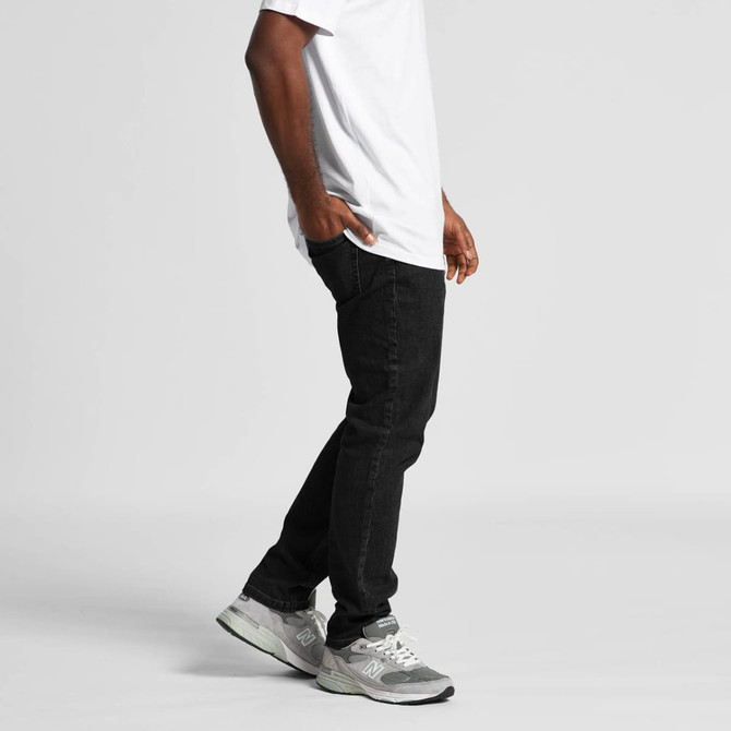 A pair of black jeans worn by a person, paired with a white t-shirt and grey sneakers. The image includes branding.