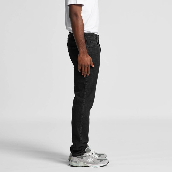 Black jeans displayed from the side, featuring a fitted design and paired with grey sneakers. There is a visible logo.