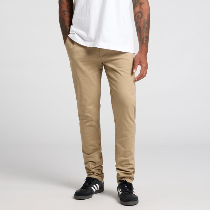 Beige pants are displayed on a model standing with hands in pockets, wearing a white t-shirt and black shoes.
