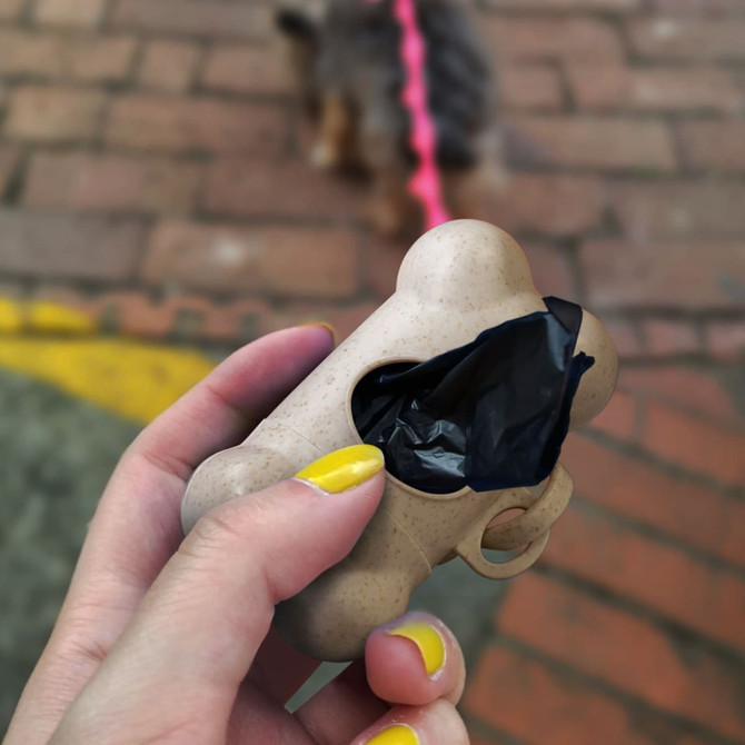 A natural-coloured pet bag dispenser shaped like a bone, holding a black waste bag, with a logo visible.