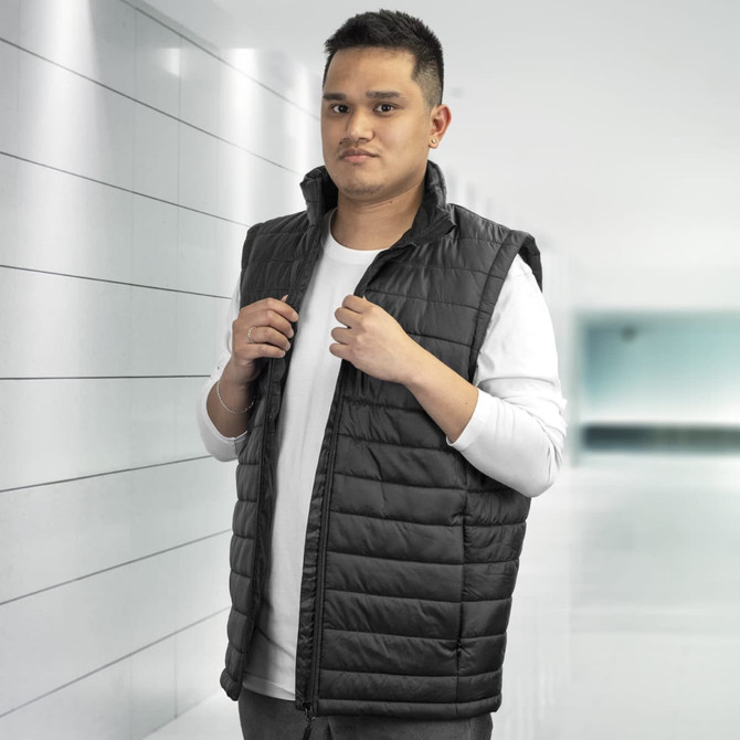 A man wearing a black puffer vest stands against a minimalist background. The vest has a zip and no sleeves.