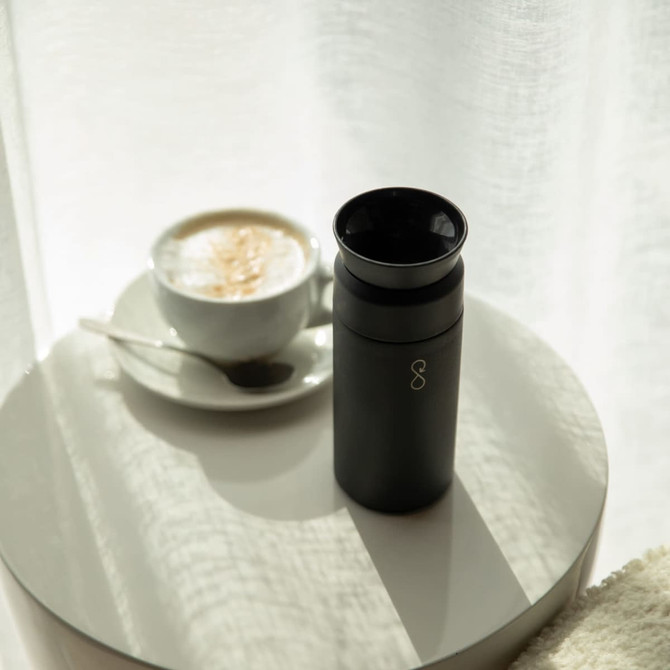 A drink bottle in matte black next to a cup of coffee on a reflective table, with soft natural light.