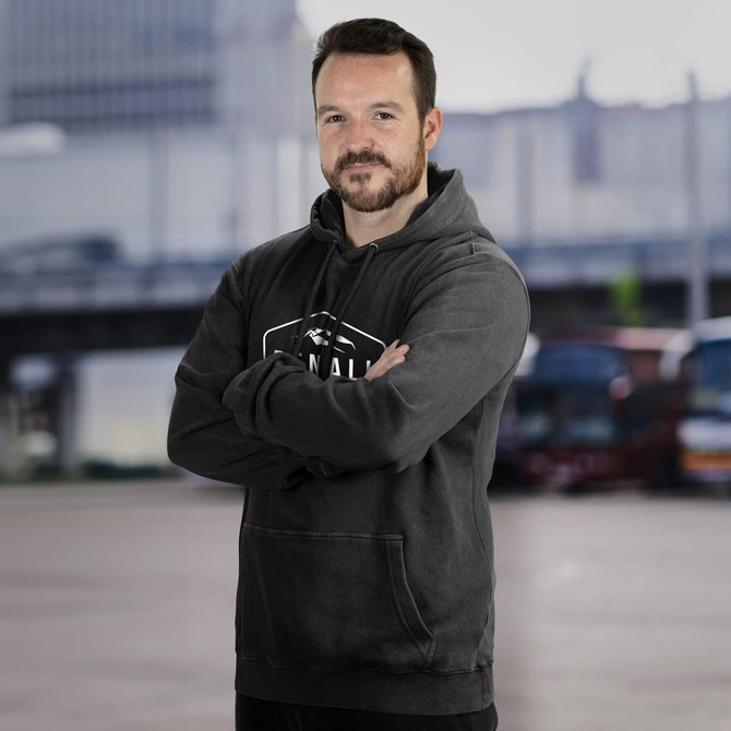 A faded grey unisex hoodie with a logo, worn by a man posing with arms crossed in an urban setting.