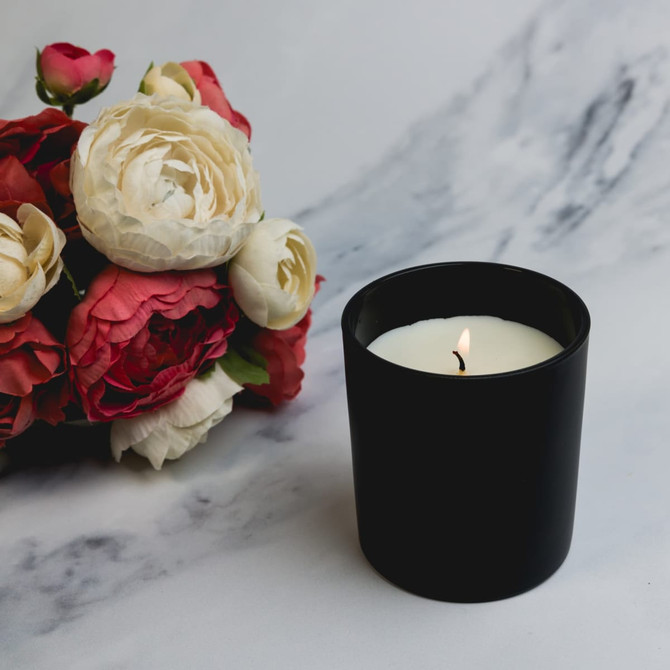 A medium-sized black glass candle with a lit wick, accompanied by a bouquet of faux flowers in shades of red and white.