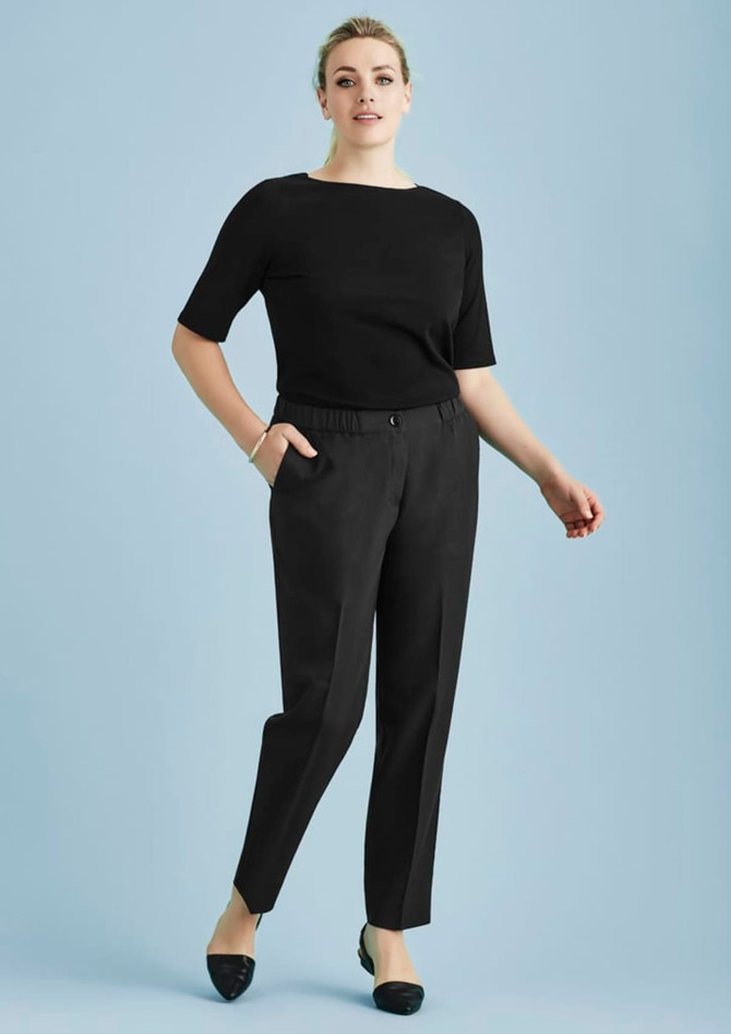 A woman in a black top and black ultra comfort waist pants poses against a light blue background. The pants feature a waistband and pockets.