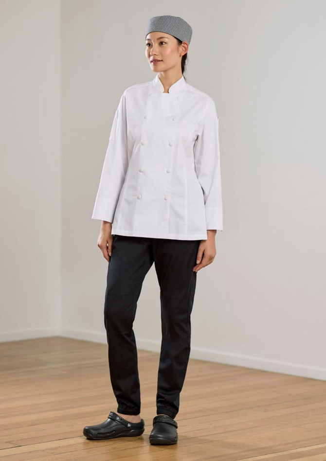 A woman in a white chef jacket and black jogger pants stands against a neutral background. She wears a grey checked head covering and clogs.