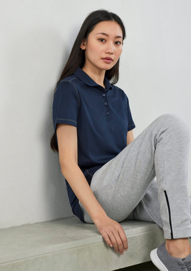 A woman wearing a navy short sleeve polo shirt sits on a bench, paired with grey athletic pants. The shirt features a logo.