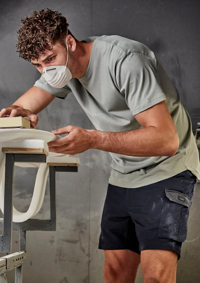 A man in a light green T-shirt and dark shorts, wearing a face mask, works at a table. The shirt has a logo.