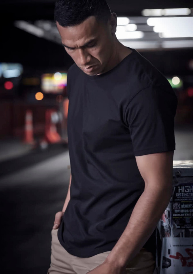 A black men's t-shirt is displayed on a model, set against a blurred urban background with street signs.