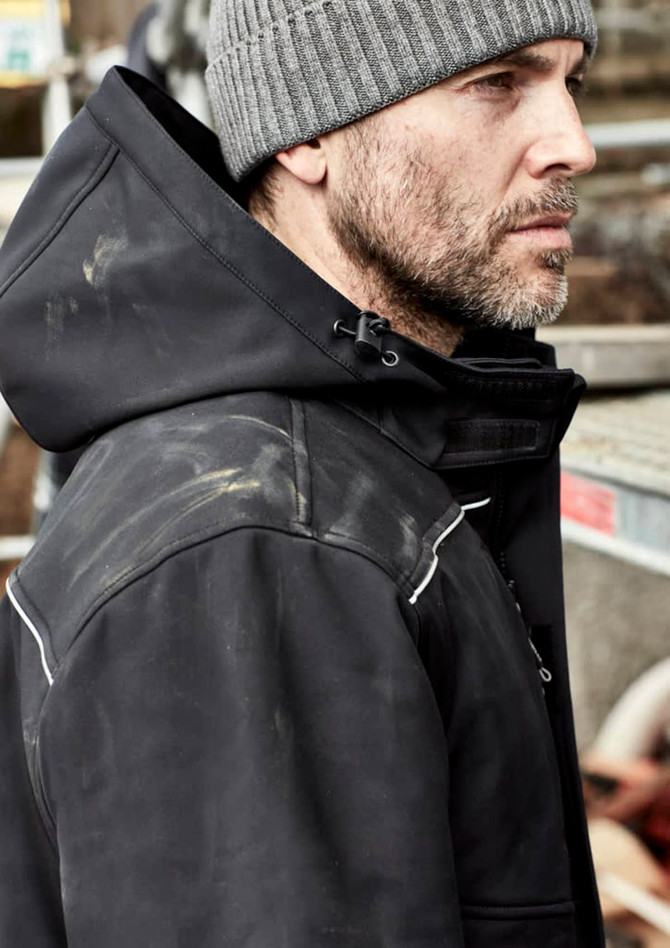 A black unisex softshell jacket with a hood, worn by a man with a grey beanie, features a logo and some dirt stains.