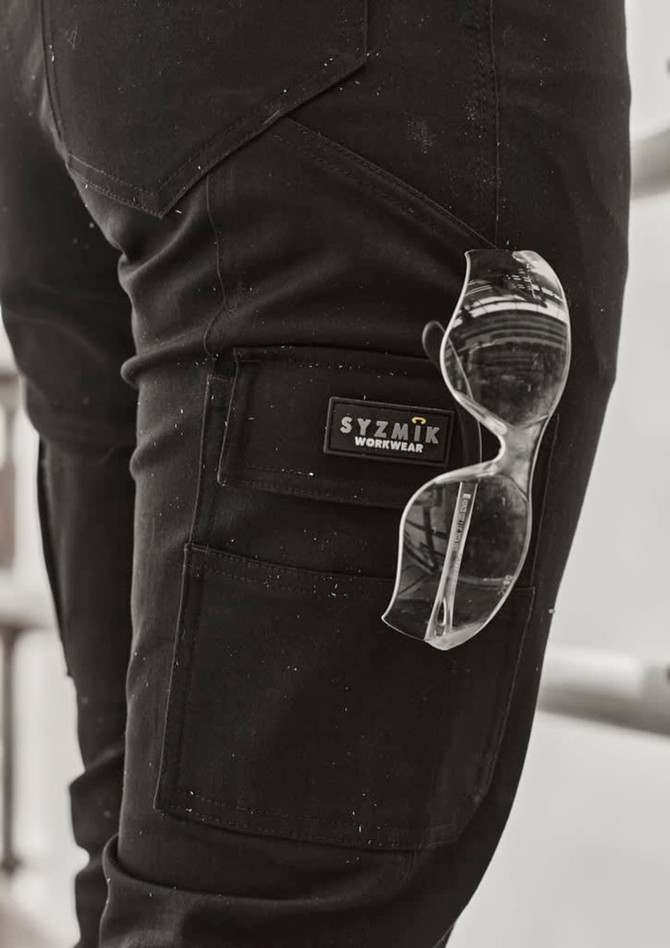 A close-up of black cargo pants with a logo and clear sunglasses tucked into the pocket.