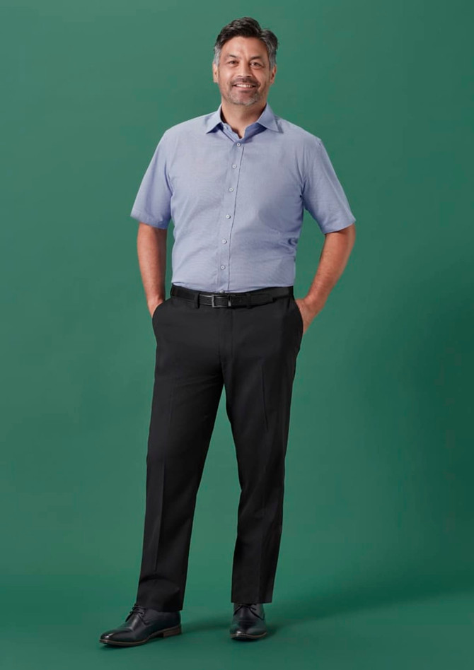 A man in a light blue short-sleeve shirt and black adjustable waist straight leg pants stands against a green backdrop.