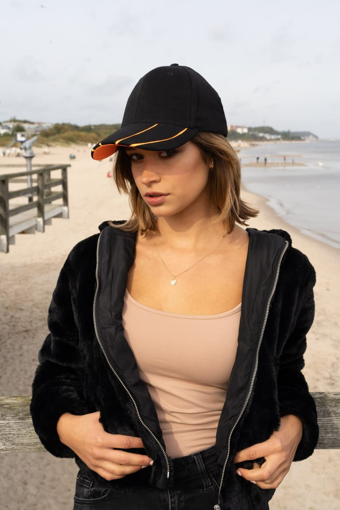 A black cap with a hi-vis orange peak worn by a woman on a beach, in a casual outfit and standing near a boardwalk.