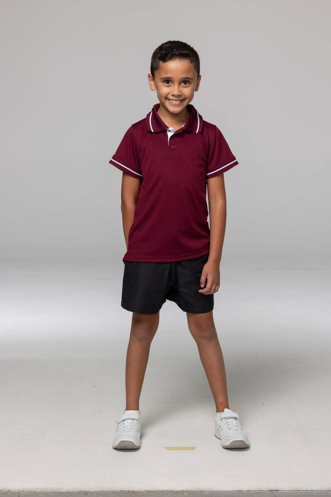 A boy wearing a maroon polo shirt with white trim, paired with black shorts and white sneakers. The shirt has a logo.