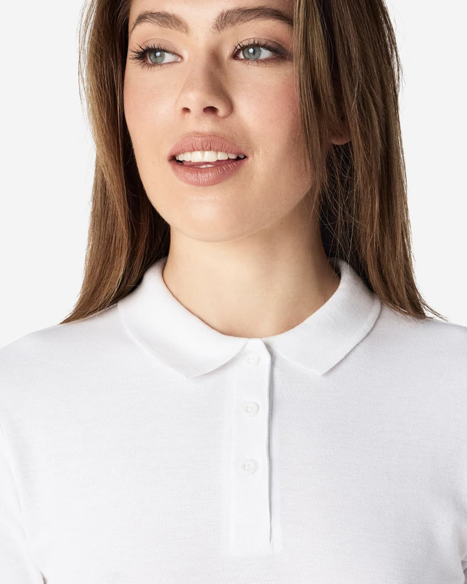 A woman wearing a white Gildan Softstyle Ladies' Double Pique Polo with a classic collar. The polo features a logo.