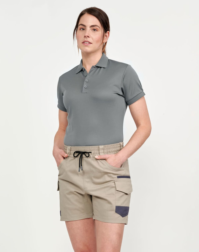 A woman wearing unisex cotton stretch drill shorts in beige, paired with a grey polo shirt. The shorts feature pockets and a drawstring.