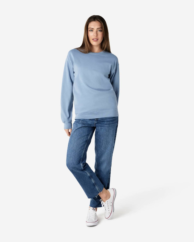 A woman stands wearing a light blue Gildan Softstyle sweatshirt paired with blue jeans and white sneakers.