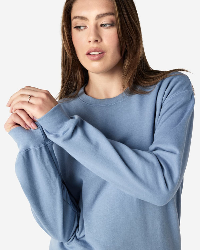 A blue Gildan Softstyle adult sweatshirt is worn by a model adjusting their sleeves, featuring a logo on the garment.