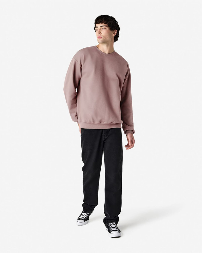 A male model wears a dusty pink Gildan Softstyle Adult Sweatshirt with black pants and black sneakers. The sweatshirt has a simple design.
