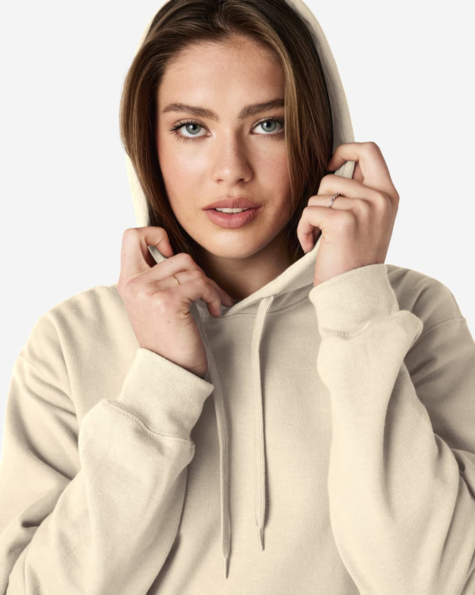 A model wearing a beige Gildan Softstyle adult hooded sweatshirt with the hoodie drawn over her head.