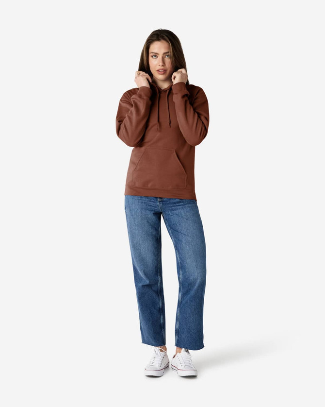 A woman wearing a brown Gildan Softstyle Adult Hooded Sweatshirt and blue jeans, standing against a plain background.