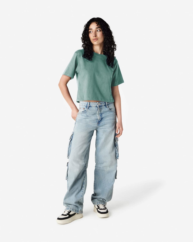 A model wears a green boxy t-shirt with light blue cargo jeans and sneakers, featuring a casual style.