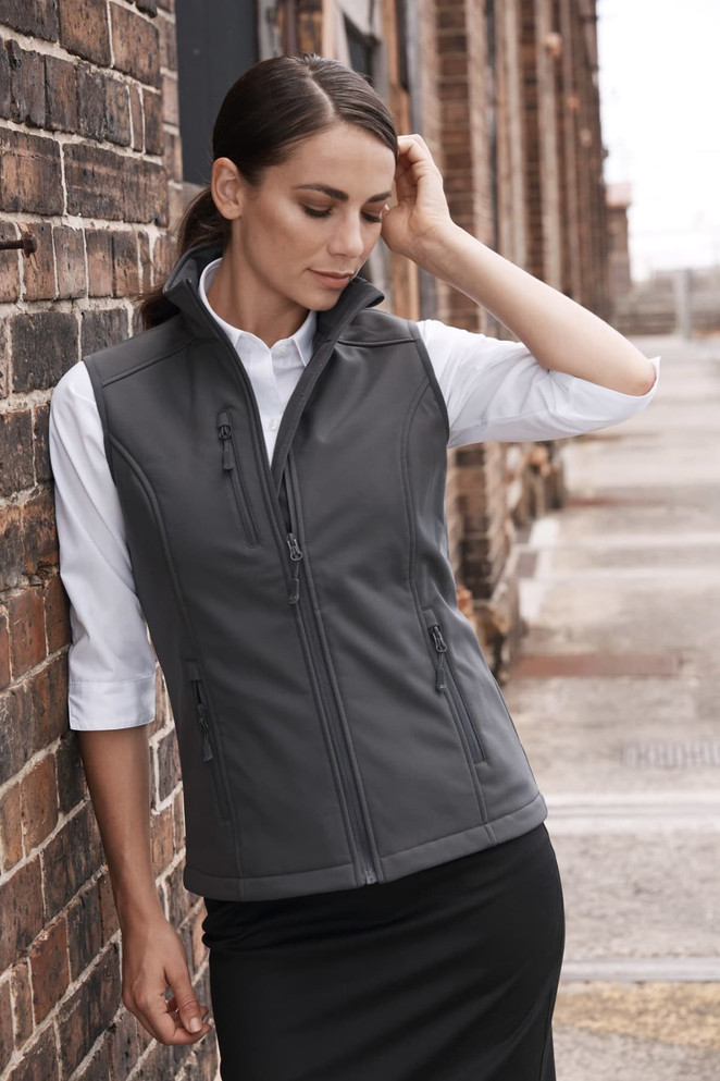 A woman in a fitted grey work vest over a white shirt stands against a brick wall. The vest has a logo on it.