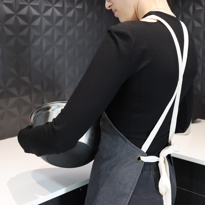 A woman wearing a grey canvas apron with white straps holds a metal mixing bowl in a kitchen setting.
