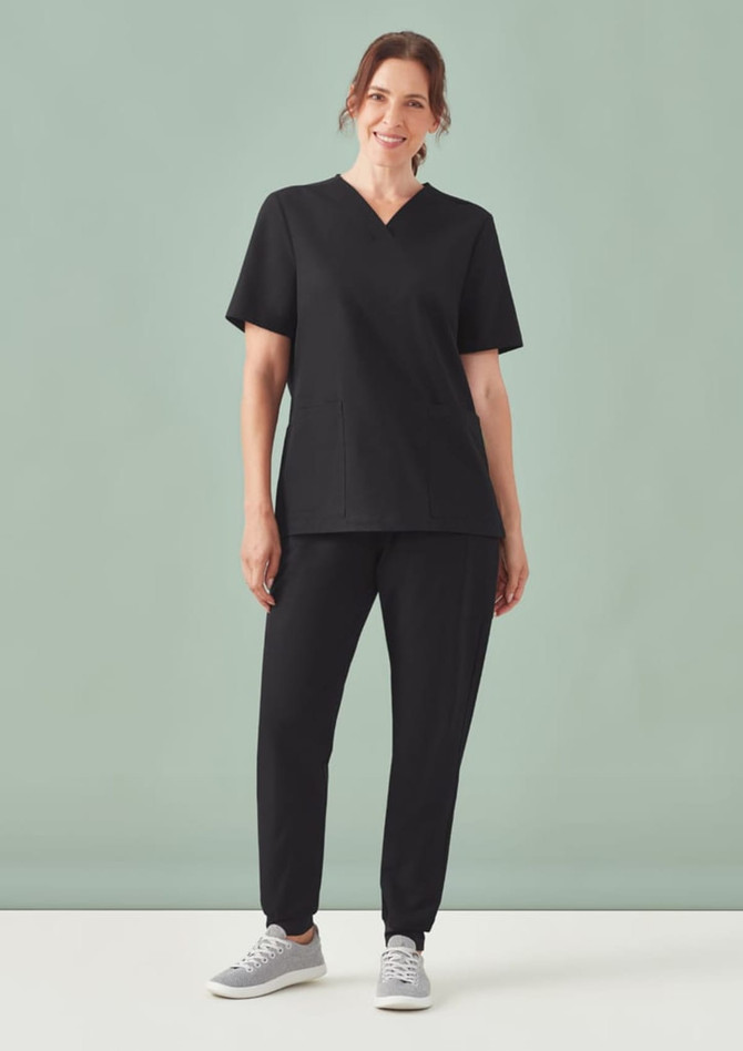 A woman stands wearing a black V-neck scrub top and matching black scrub pants, paired with grey sneakers.