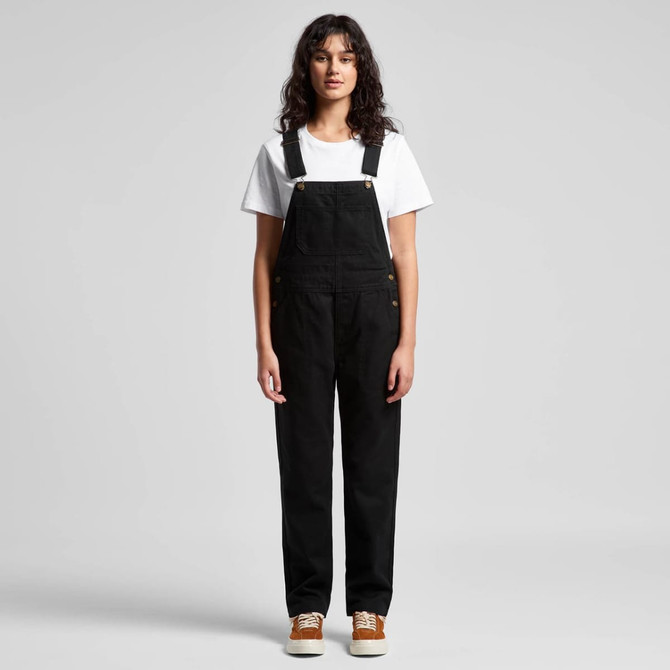 A pair of black canvas overalls worn by a model, paired with a white t-shirt and brown shoes.
