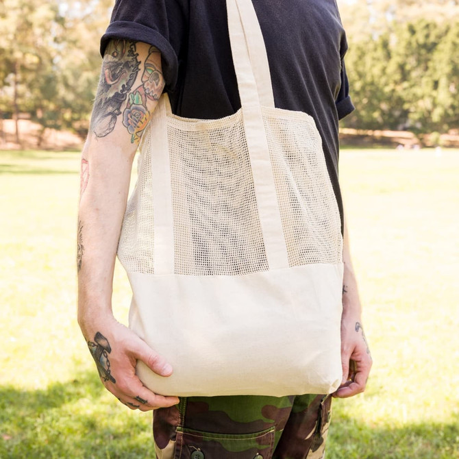 A cream-coloured mesh tote bag with long handles, held by a person in a casual outfit.