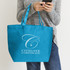 A blue tote bag with a logo, held by a person wearing a grey sweater and jeans, holding a mobile phone.