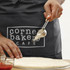 A dark grey waist apron features a logo, with a person using a scoop above a pizza dough.