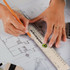A 30cm ruler with a logo, being held over architectural plans while a hand uses a pencil. The ruler is a transparent colour with metal edges.