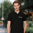 A black men's polo shirt featuring a logo on the chest, presented against an outdoor background.
