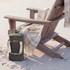 A wine cooler bag in olive green with a shoulder strap rests on the sand beside a wooden chair and a blanket.