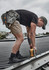 A person wearing khaki cargo shorts and a black t-shirt is using a power drill on a roof. Tools hang from a tool belt.