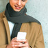 A grey cable knit scarf is worn by a person in a tan coat, holding a smartphone against a textured background.