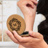 A cork compact mirror held in a hand, featuring a decorative design on the lid, set against a soft background.