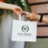 A medium flat handle paper bag in white, featuring a branding logo, held by two hands.