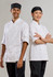 Two chefs stand back-to-back, both wearing white long sleeve chef jackets and black trousers, with one wearing a chef hat and the other a head scarf.