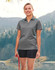 A woman stands outdoors wearing a grey polo shirt with blue contrast accents, alongside black shorts and holding a leash.