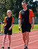 Men's TrueDry tri-colour short-sleeve pique polo in black and orange, paired with matching shorts, worn by two people on a track.