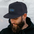 A black flat peak cap with a patch, worn by a man with a beard, against a textured background.