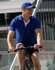 A man in a bright blue polo shirt and cap rides a bicycle, smiling. The polo features a logo.