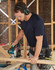 A navy blue short-sleeve work t-shirt worn by a man using a circular saw at a construction site.