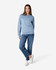 A woman stands wearing a light blue Gildan Softstyle sweatshirt paired with blue jeans and white sneakers.