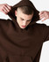 A brown unisex hooded sweatshirt with a front pocket and a model adjusting the hood. It features a logo.