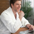 A man wearing a white waffle bathrobe sits at a table with a laptop and a cup, gazing thoughtfully.